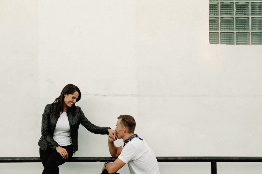 A woman in a leather jacket sits on a railing talking with a man in a white tee against a white wall.