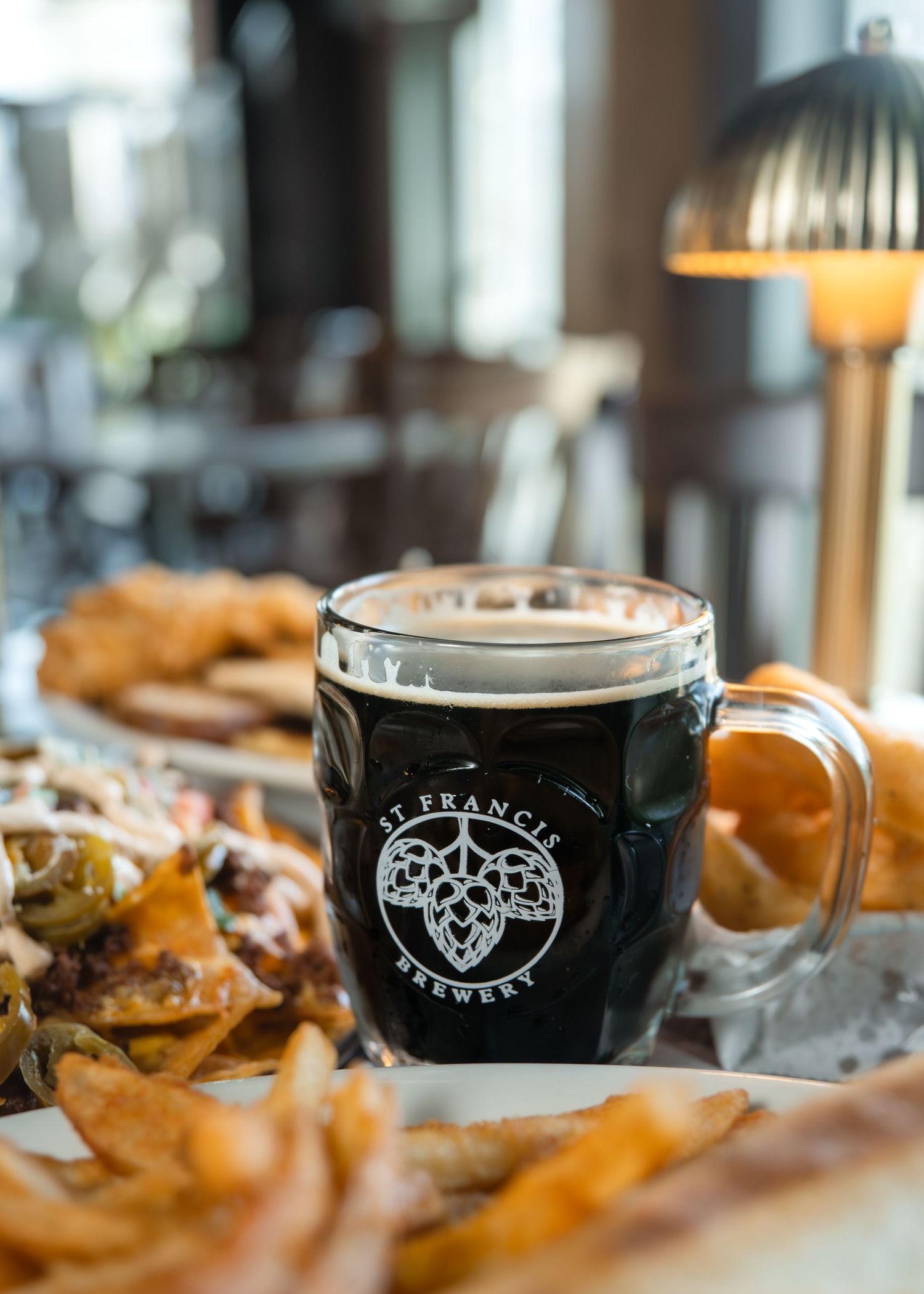 A dark beer in a St. Francis Brewery glass mug served alongside fries and appetizers on a restaurant table.