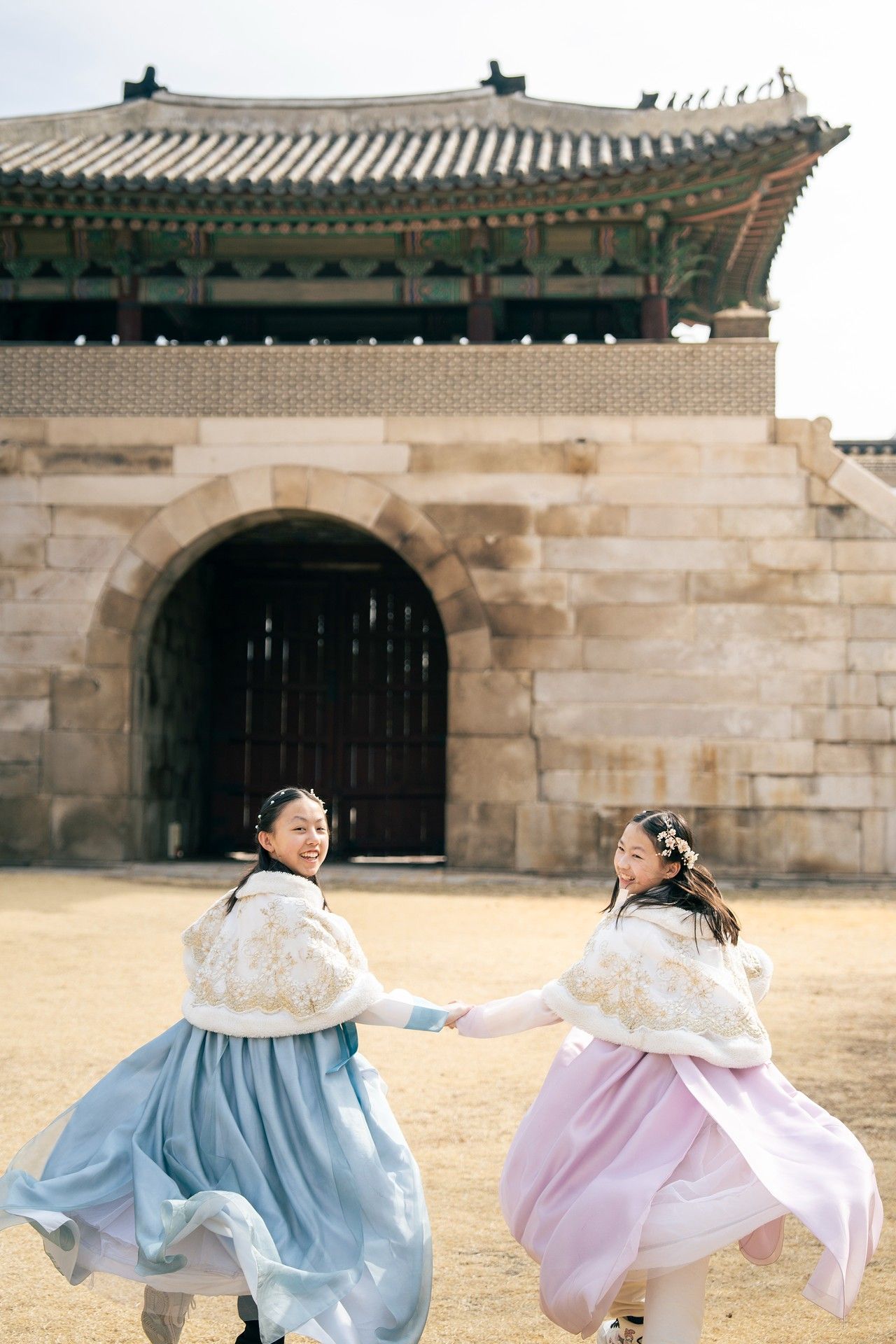 Two women in traditional Korean hanbok dresses hold hands and spin joyfully outside a historic palace gate in Seoul.