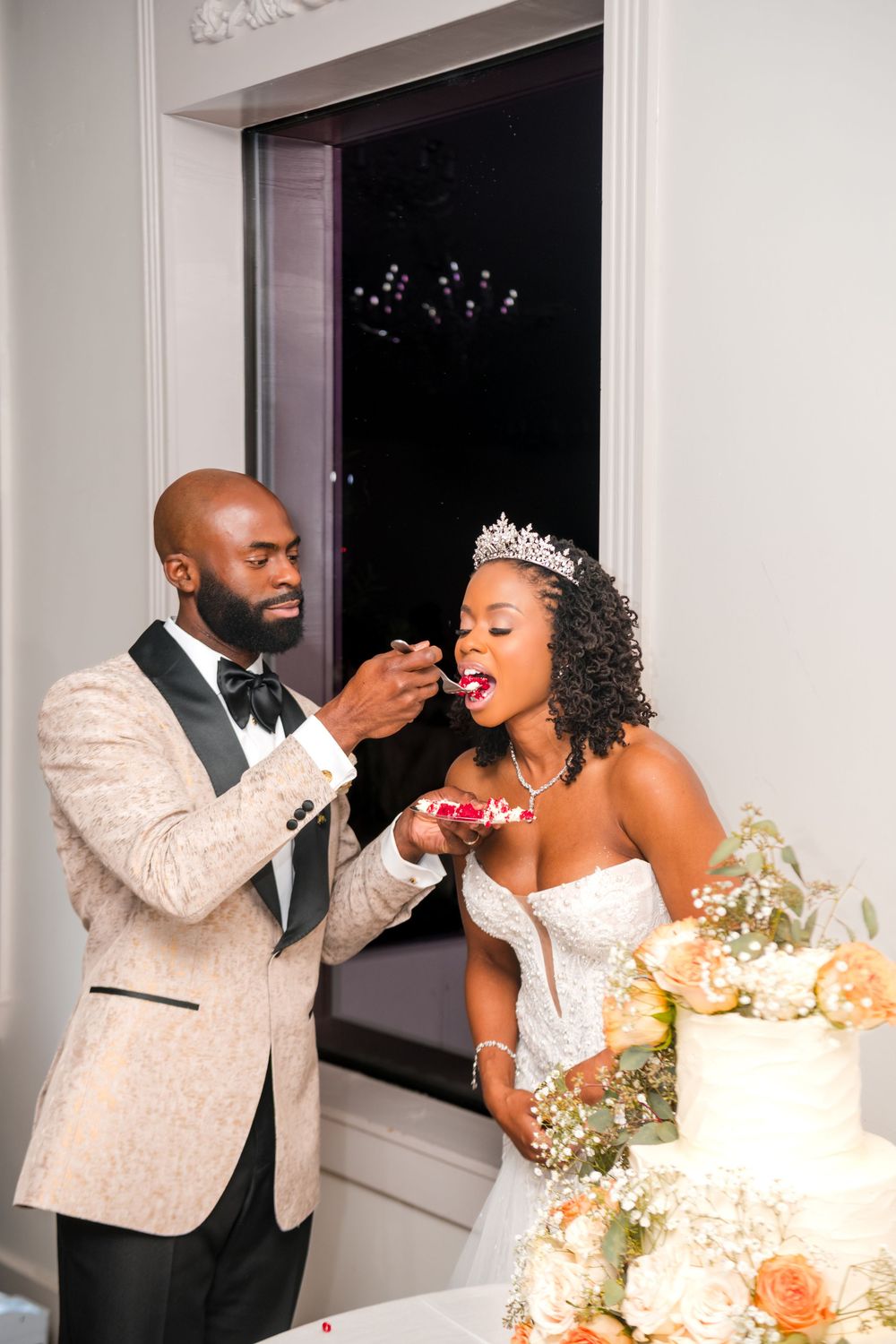 Wedding couple sharing cake during reception in an elegant white room with window views.