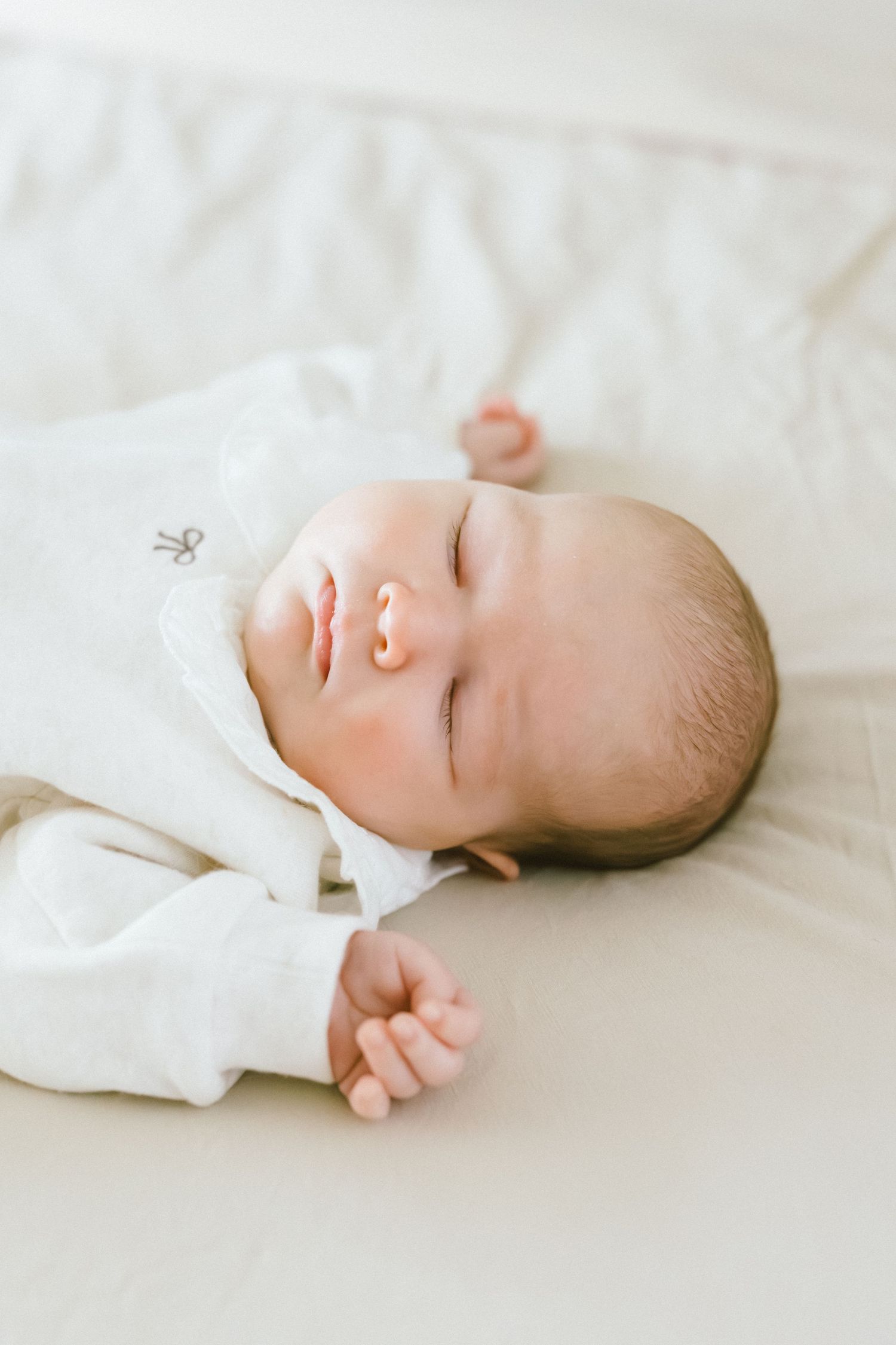 Peaceful newborn baby sleeping on white bedding wearing a white onesie with a pacifier.