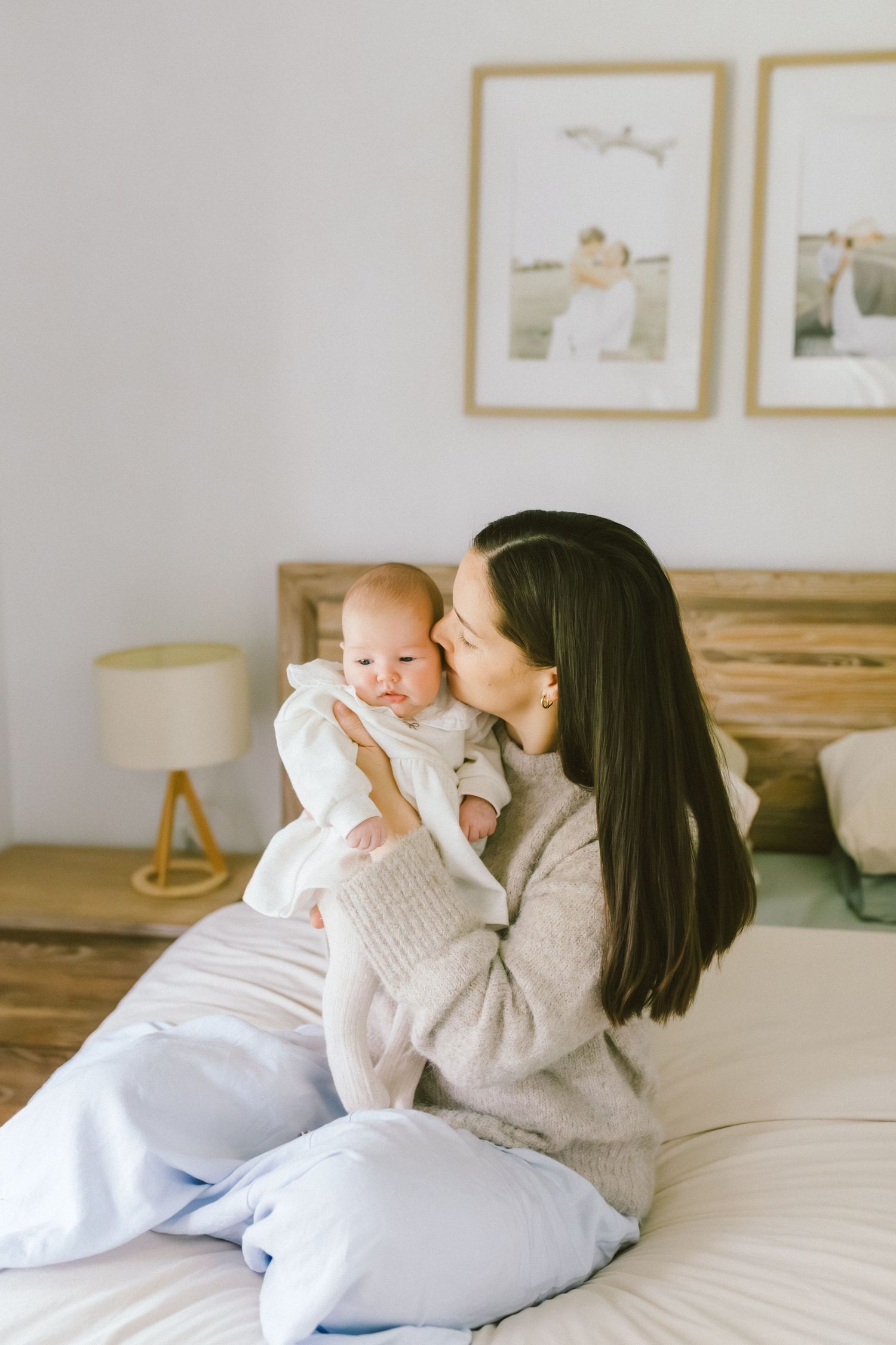 A mother sitting on a bed kisses her newborn baby in a cozy bedroom with framed photos on the wall.