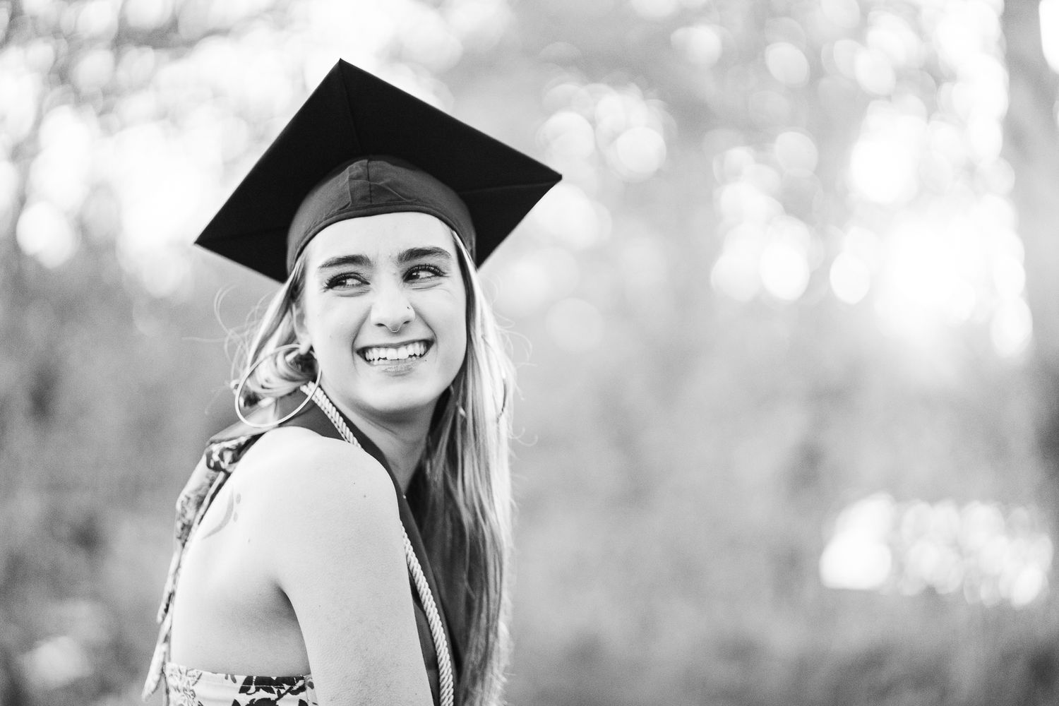 Smiling young woman wearing a graduation cap poses outdoors in a black and white portrait photo.