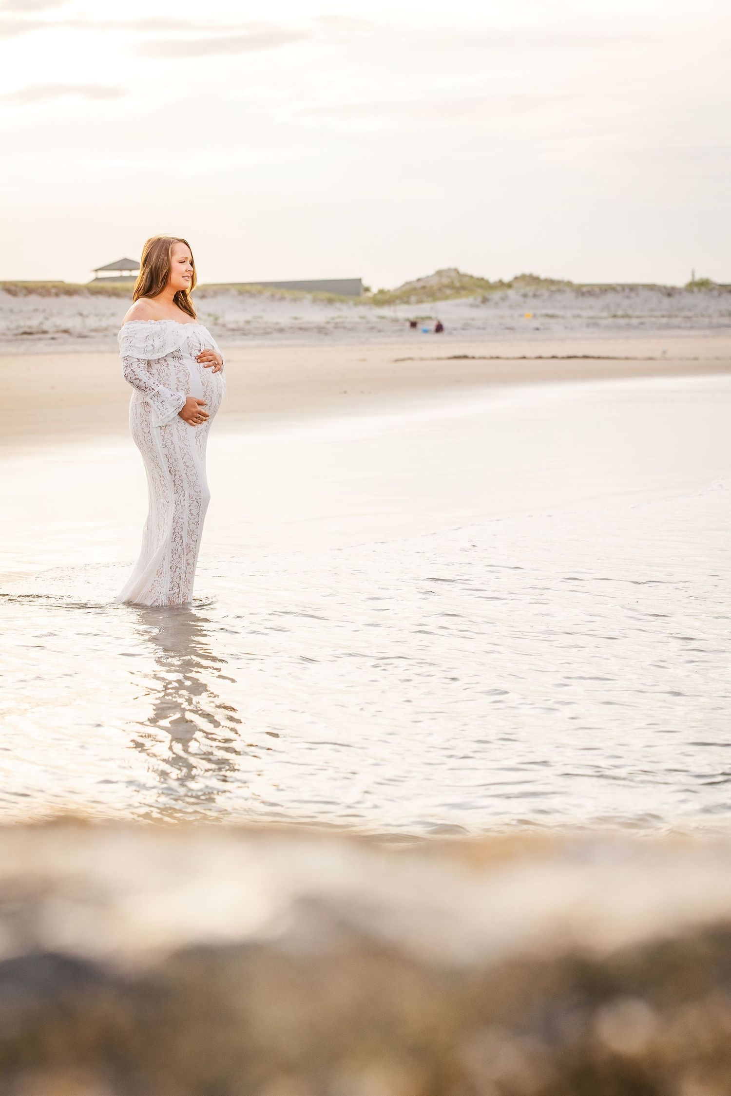 Pregnant woman in white lace gown standing in shallow ocean water at sunset on a serene beach.