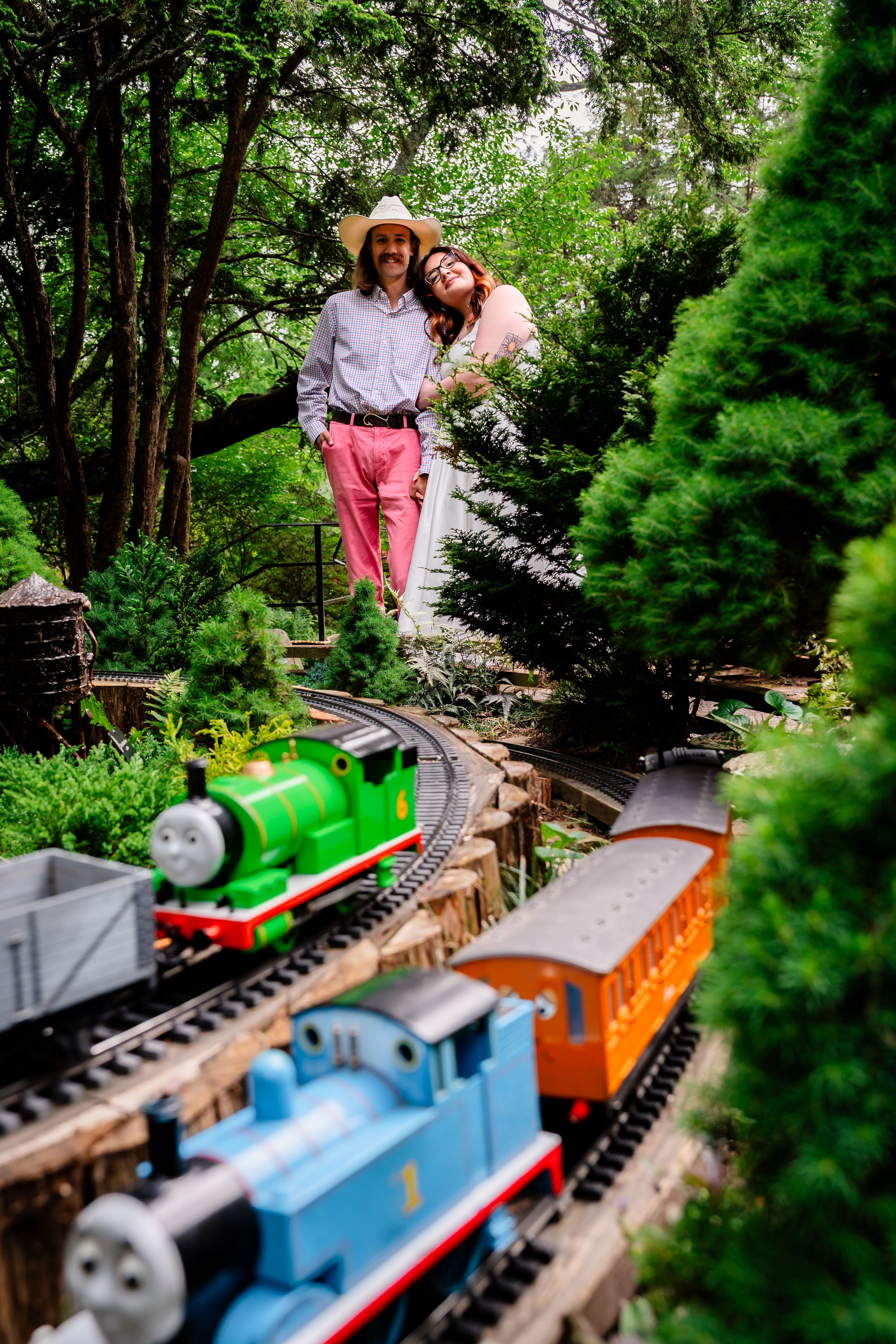 Smiling couple standing in lush garden with Thomas the Tank Engine model train set in foreground.