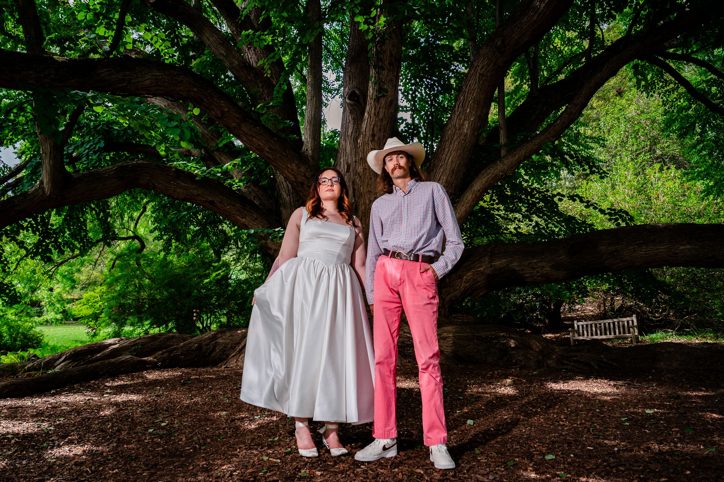 Bride in white dress and groom in pink pants posing under a large tree in a lush garden.