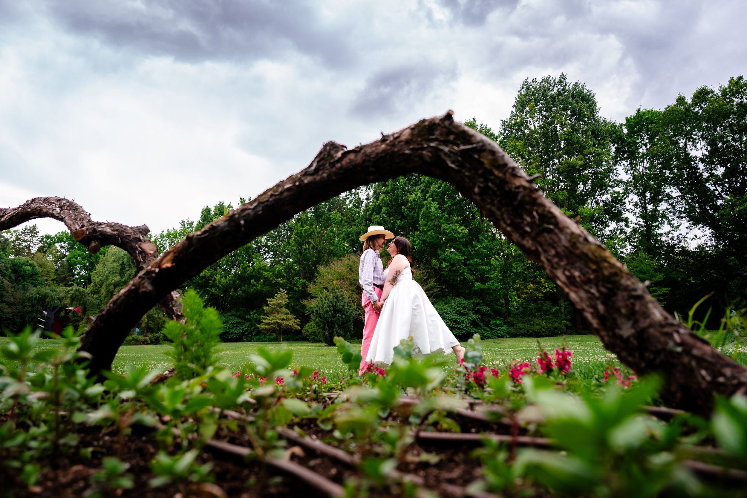 Couple kissing under arching tree branch in green garden with red flowers in foreground.