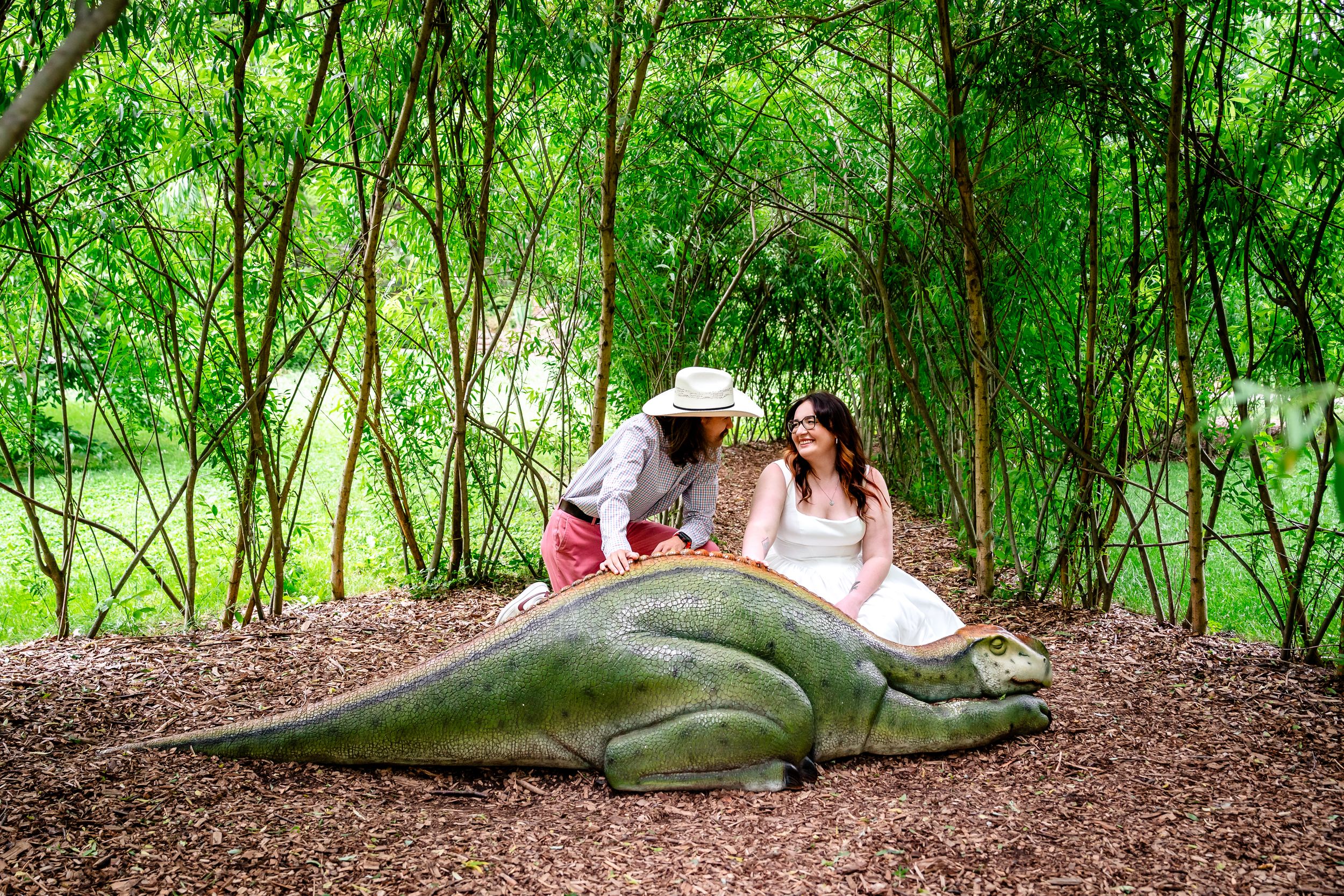 Bride and groom posing with a large dinosaur statue surrounded by bamboo in a garden.
