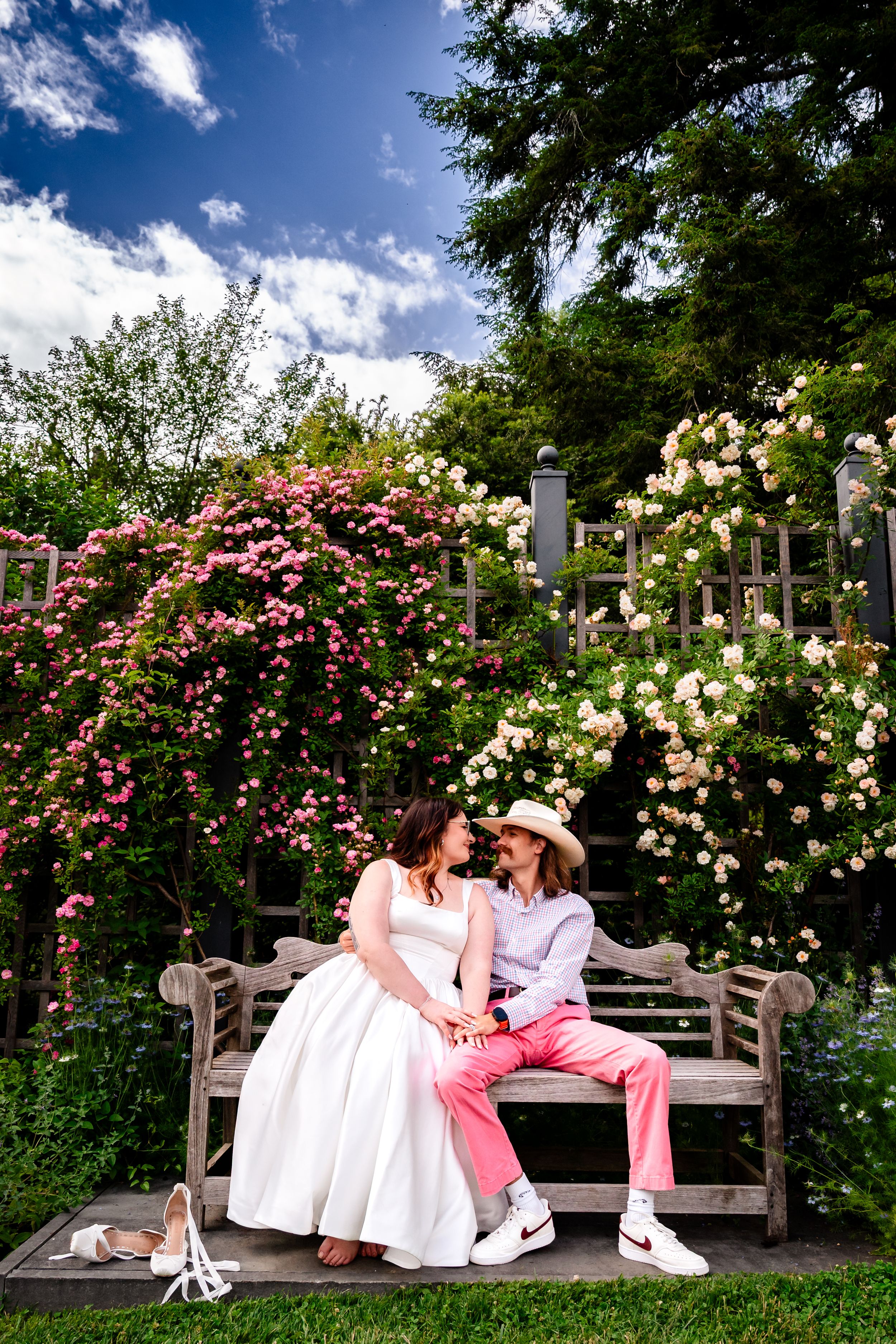 Bride and groom sitting close on wooden bench surrounded by blooming pink and white roses in garden.