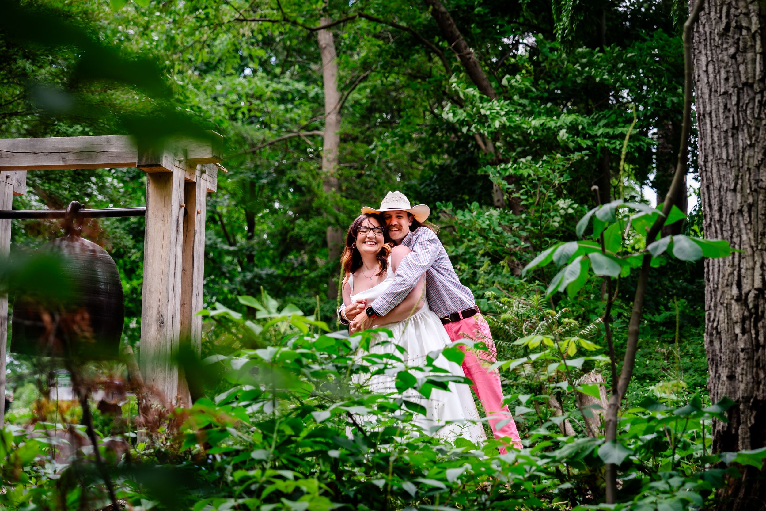 Couple embracing and smiling in a lush green garden surrounded by trees and foliage.