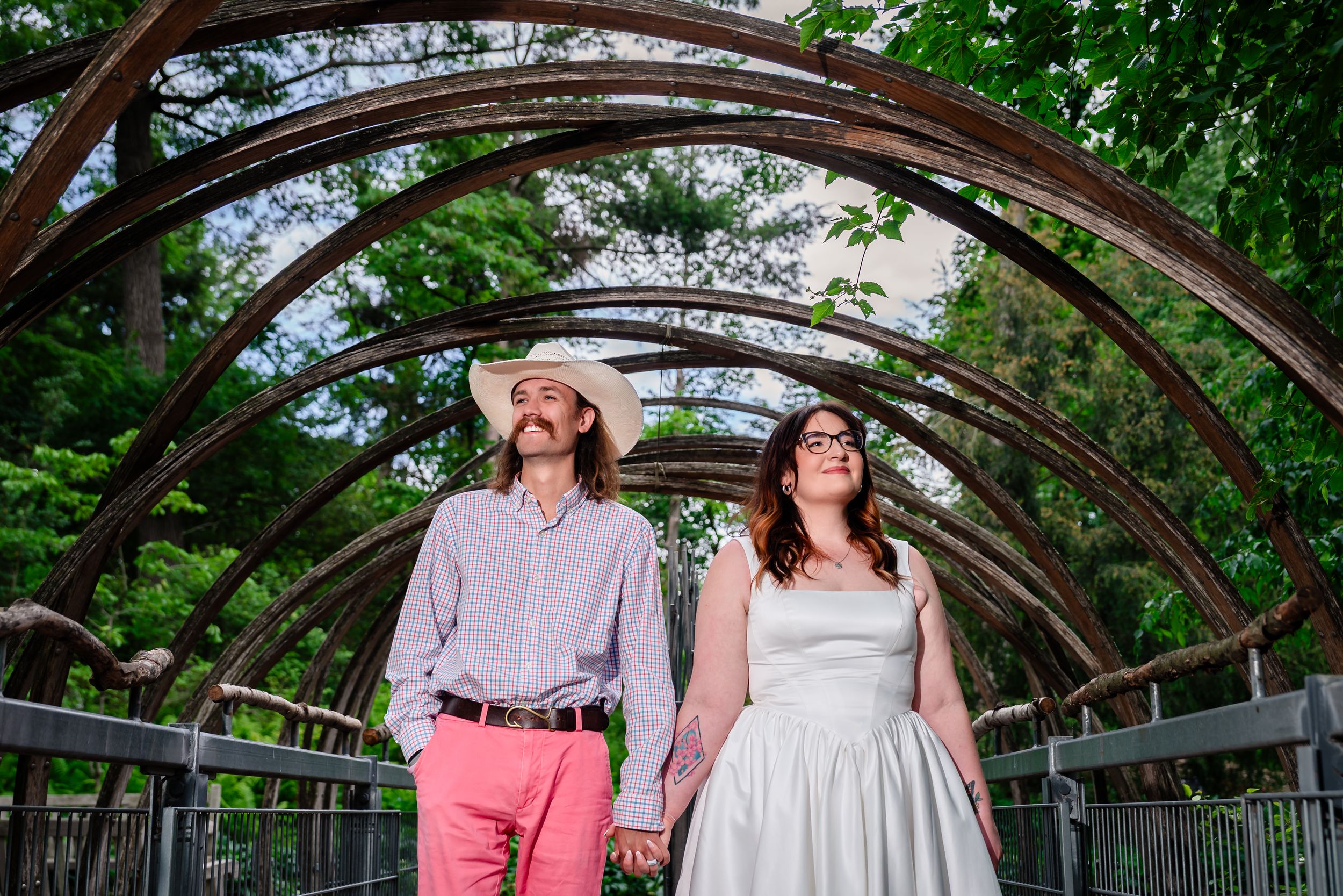 Bride and groom hold hands under rustic metal arch bridge surrounded by lush green trees on their wedding day.