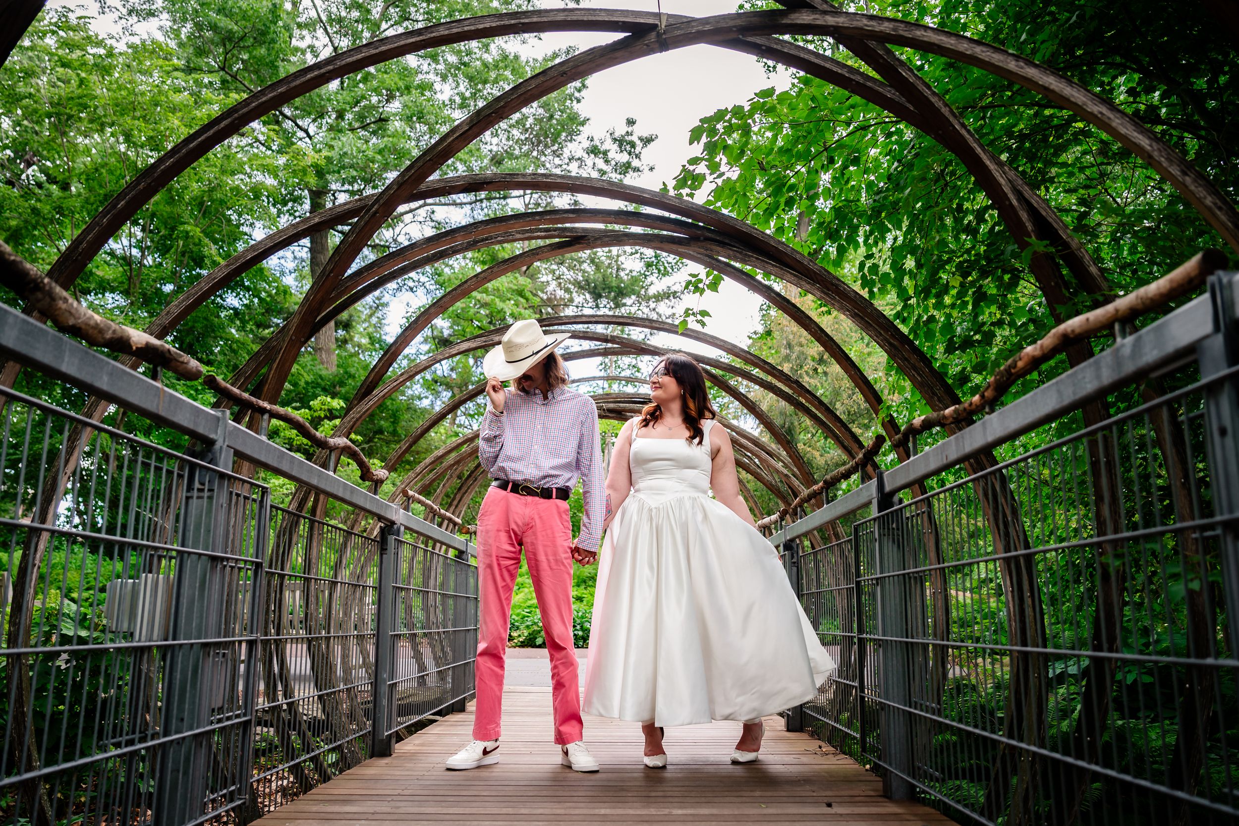 Bride and groom holding hands on a wooden bridge tunnel covered in lush green vines at an outdoor wedding venue.
