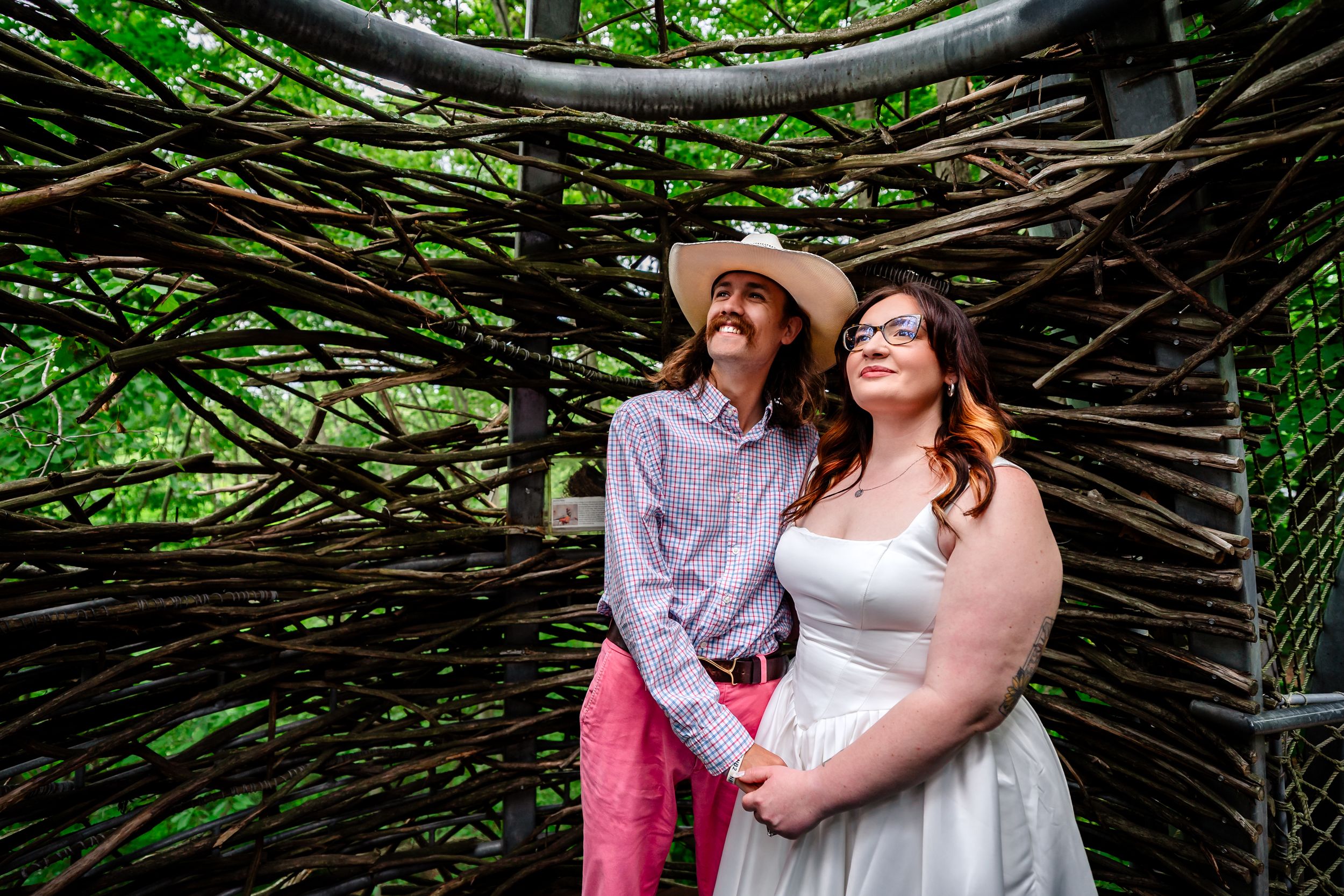 Couple in wedding attire posing under rustic woven branch arch in lush green forest setting.