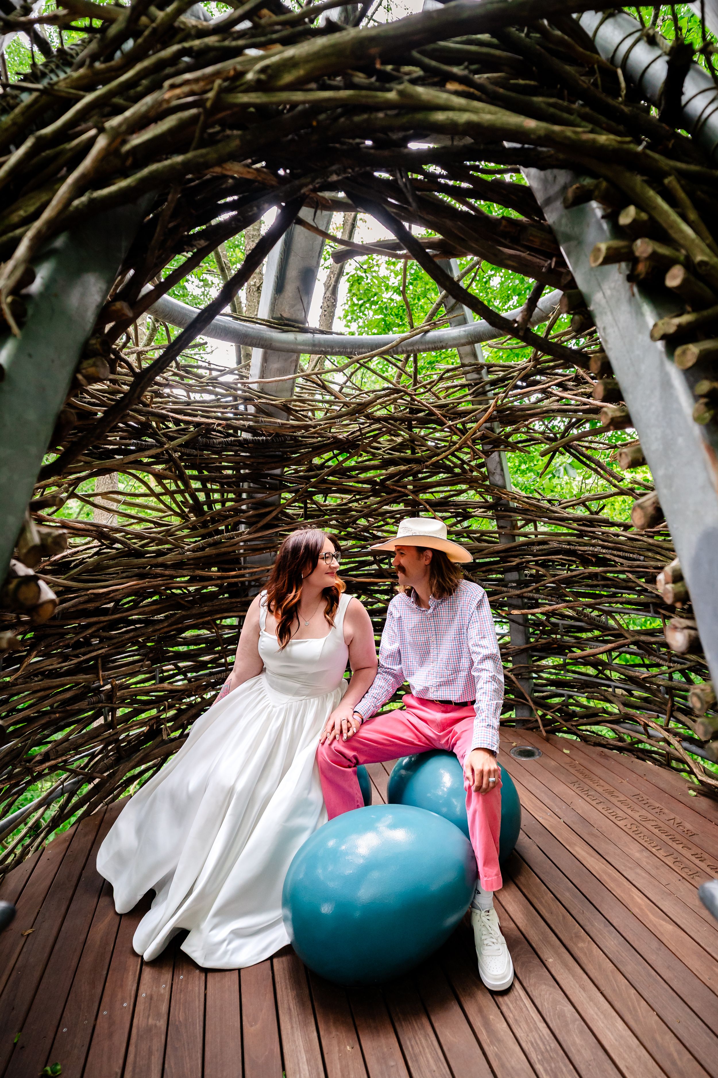Bride and groom sitting together inside a whimsical woven branch arbor on a wooden deck.