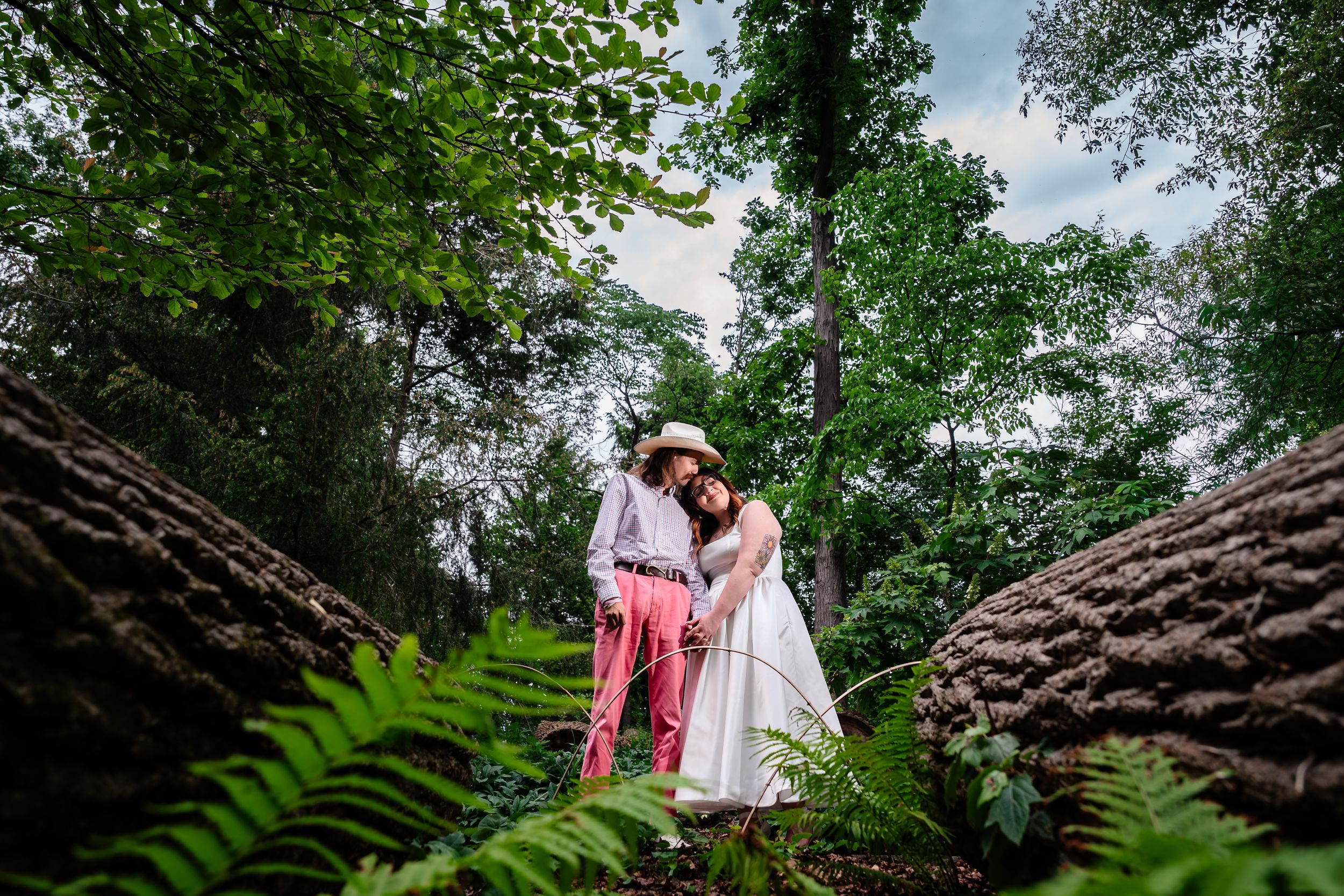 Couple in wedding attire embracing among lush green ferns and trees in a romantic forest setting.