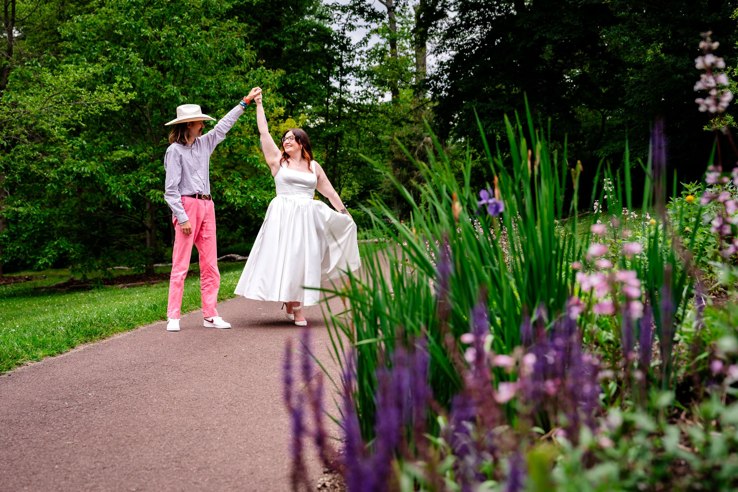 Bride in white dress dancing with partner in pink pants and sun hat on garden path surrounded by purple flowers.