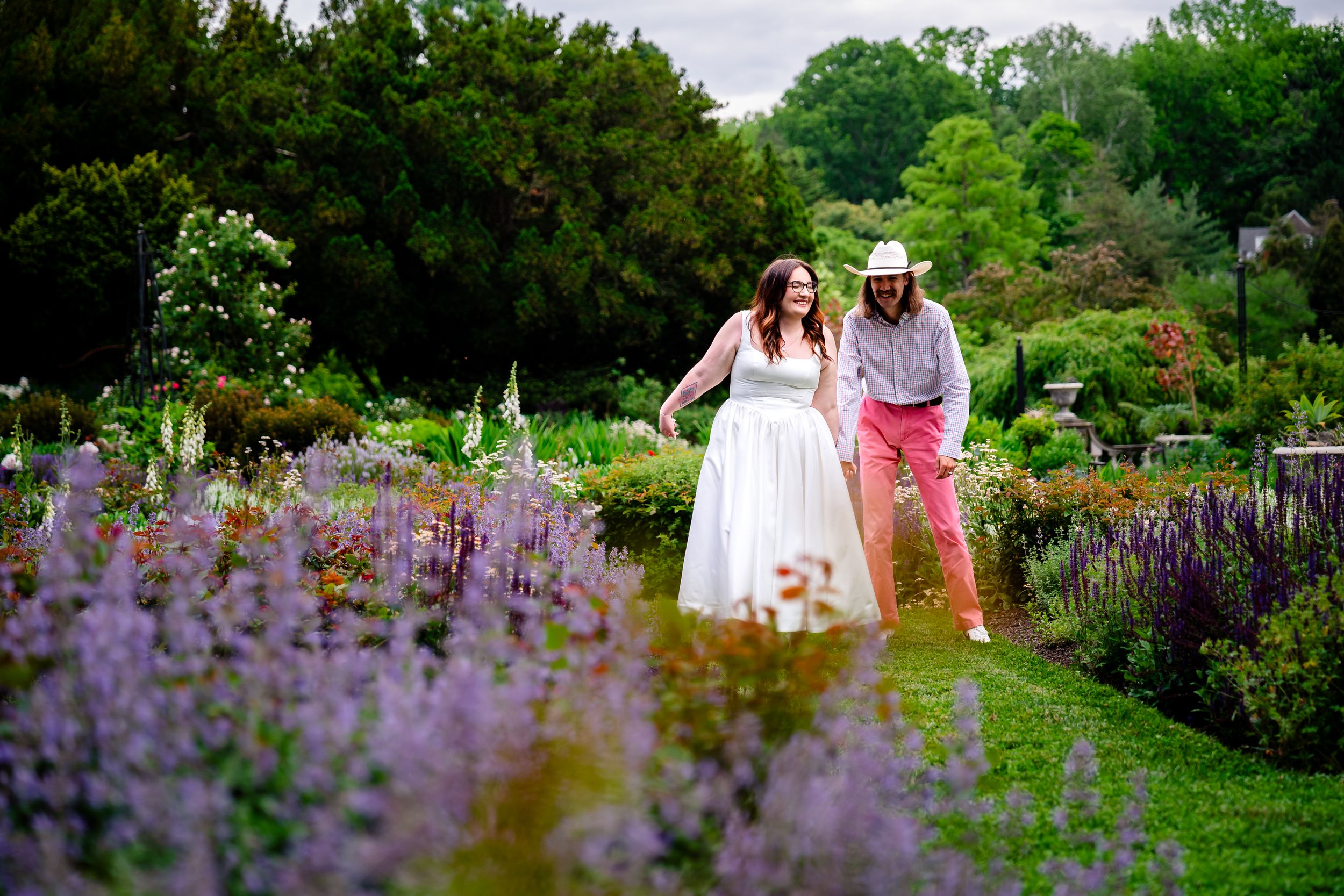 Bride and groom walking through a colorful garden with purple flowers and lush greenery on their wedding day.