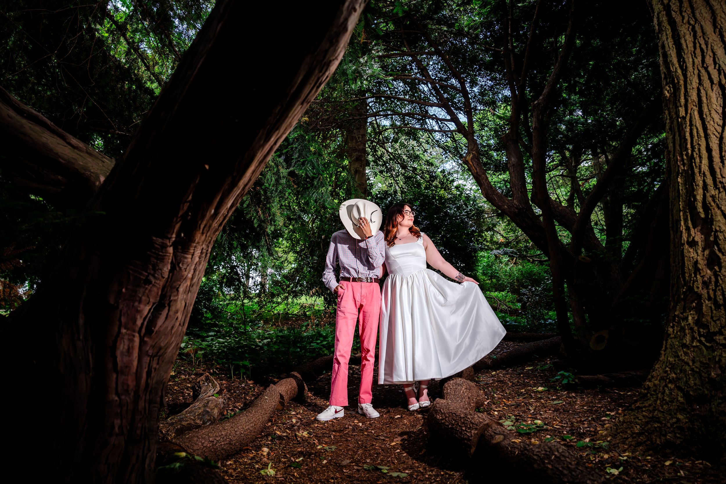 Couple in wedding attire posing romantically among twisted tree roots in a lush green forest.