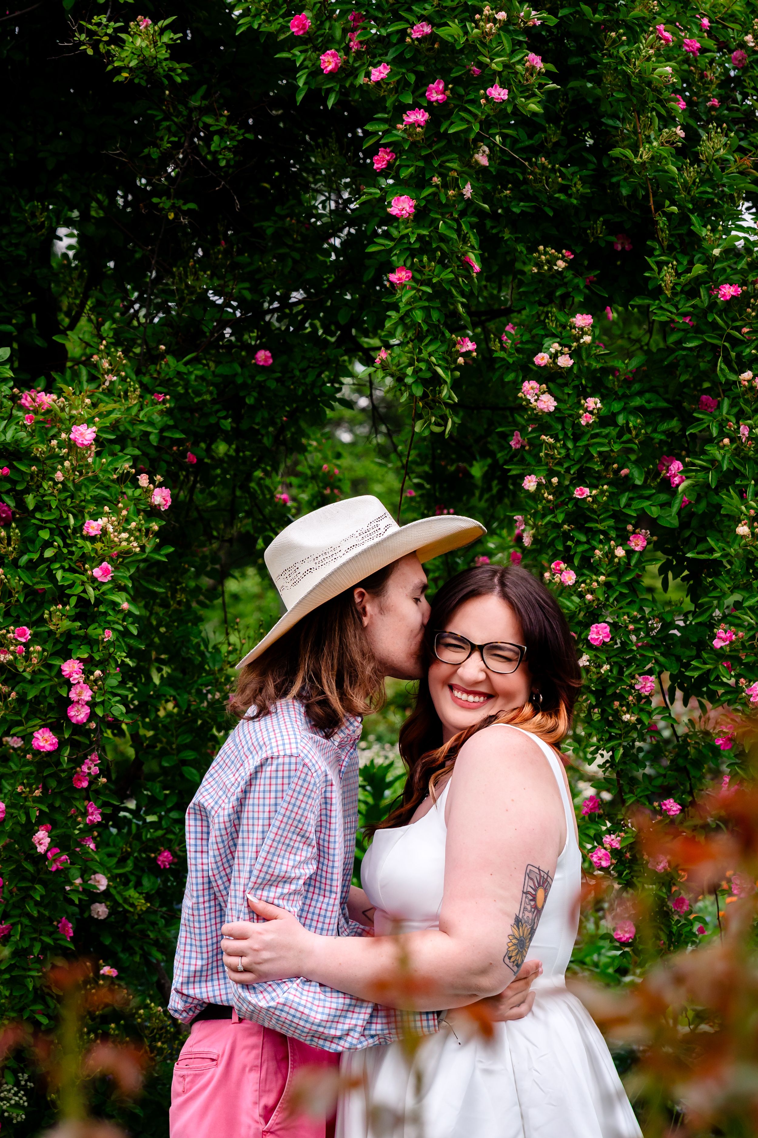 Two women embrace and kiss among pink flowering bushes in a garden, one wearing a straw hat and gingham shirt.