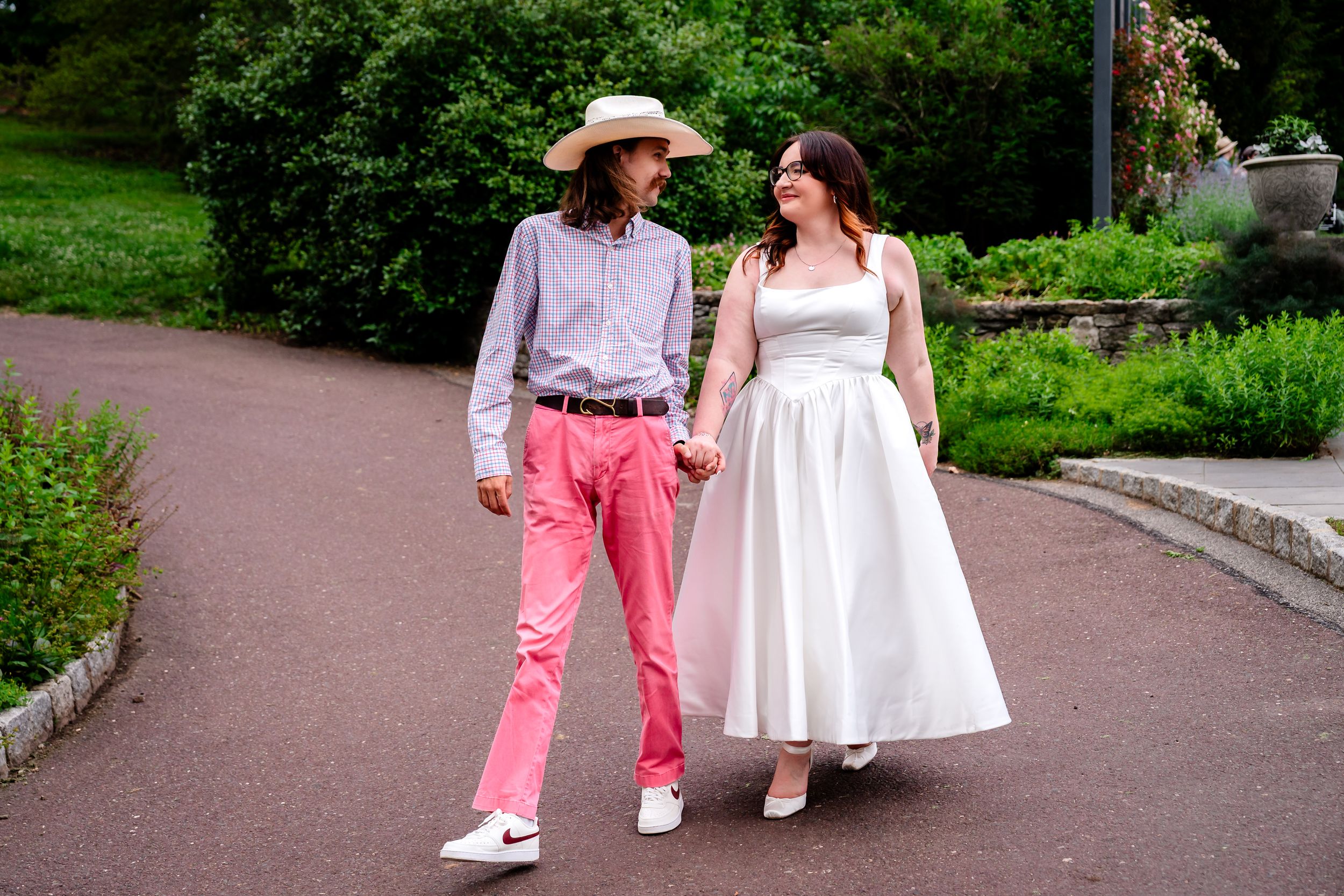 Couple holding hands walking in park, groom in pink pants and cowboy hat, bride in white dress.