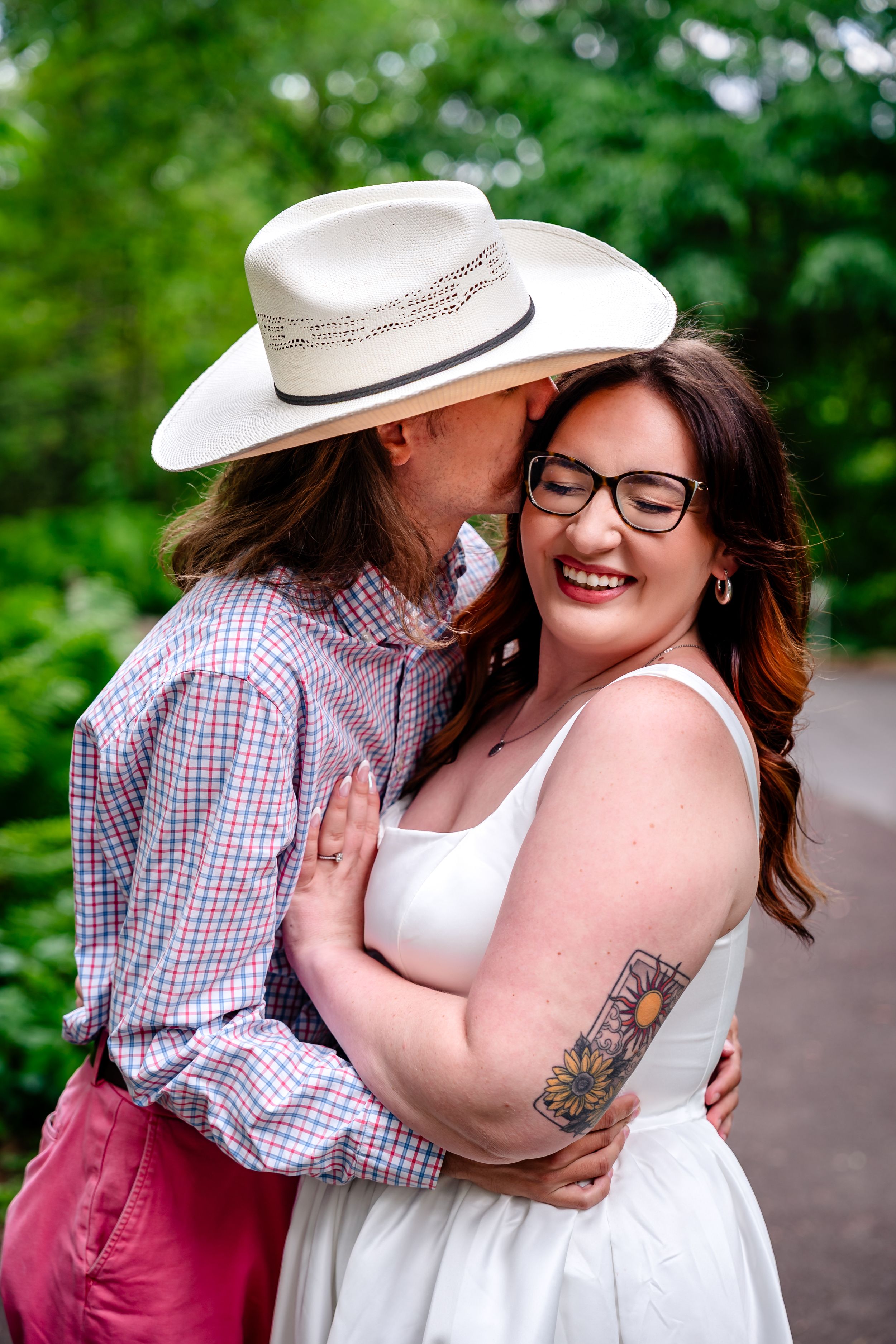 Couple embracing outdoors, man in white cowboy hat kissing woman in white dress with sunflower tattoo.