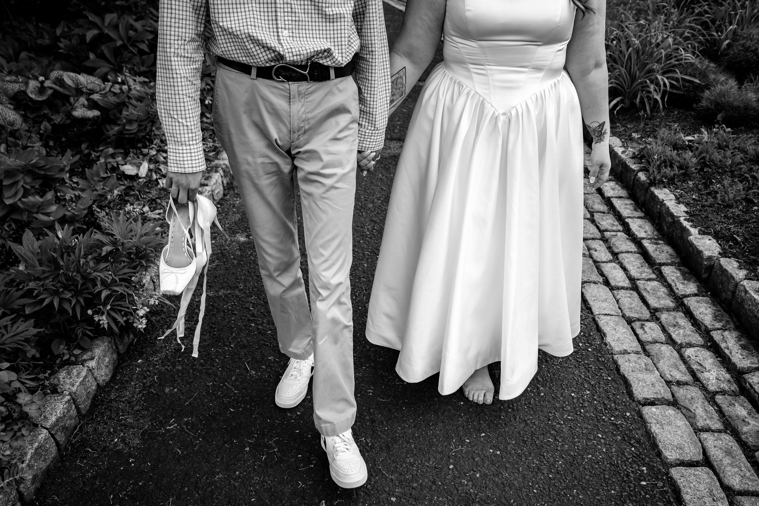 Black and white photo of a bride in white dress and groom holding heels, walking hand in hand on a cobblestone path.