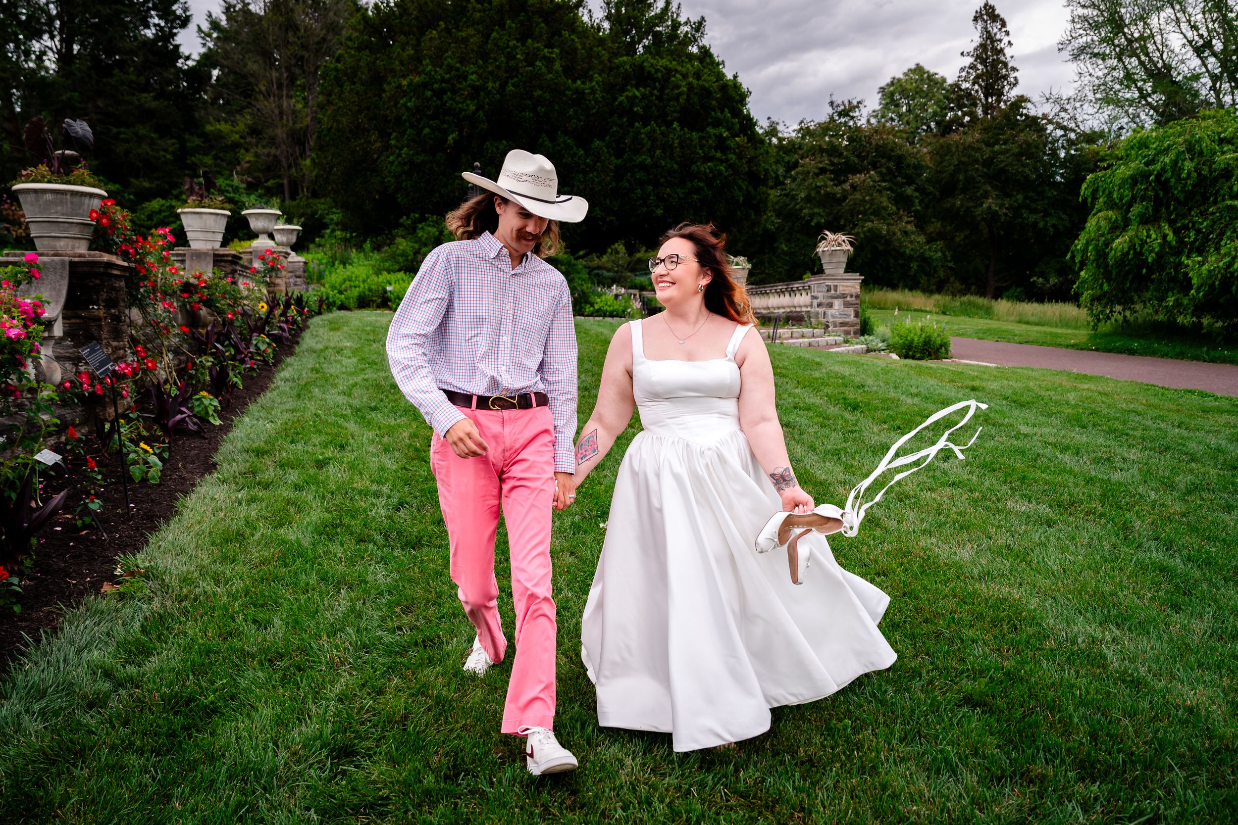 Couple walks hand-in-hand through garden, groom in pink pants and cowboy hat, bride in white dress holding flowers.