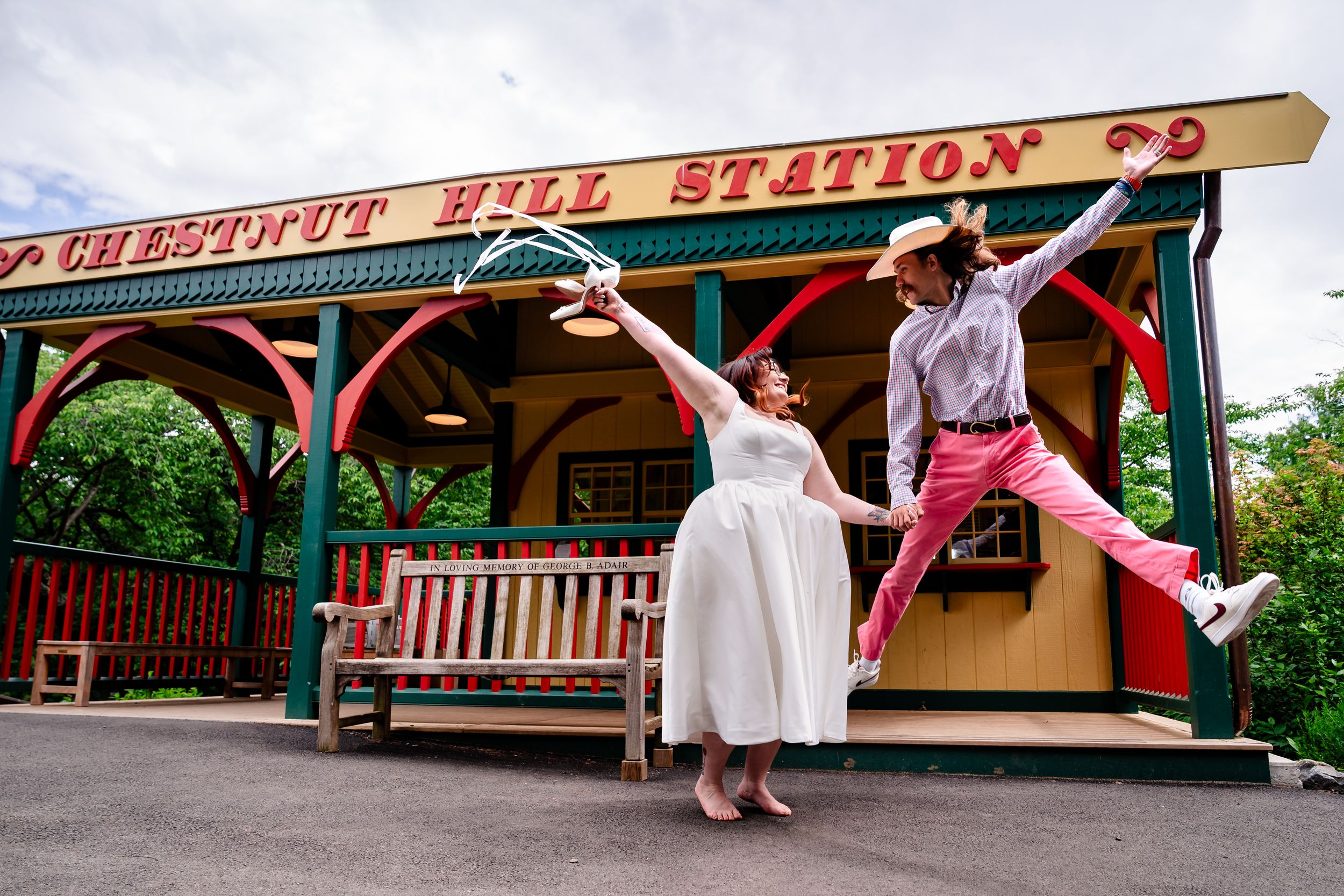 Joyful couple jumping outside Chestnut Hill Station, bride in white dress, groom in pink pants.