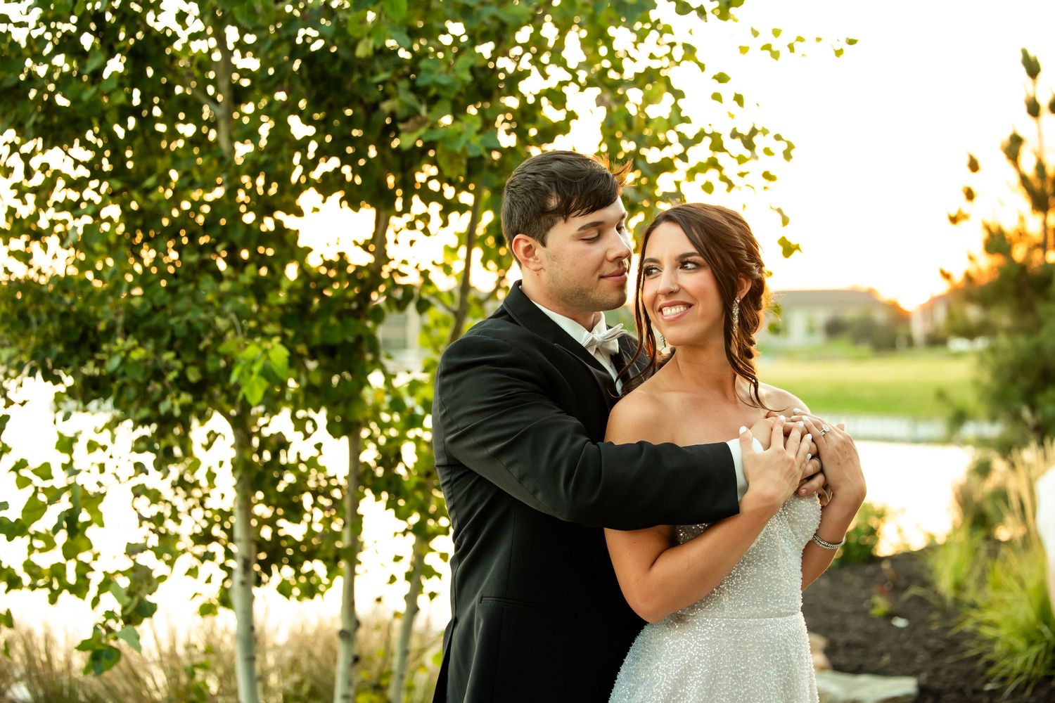 Bride and groom share tender moment outdoors surrounded by lush green trees at sunset.