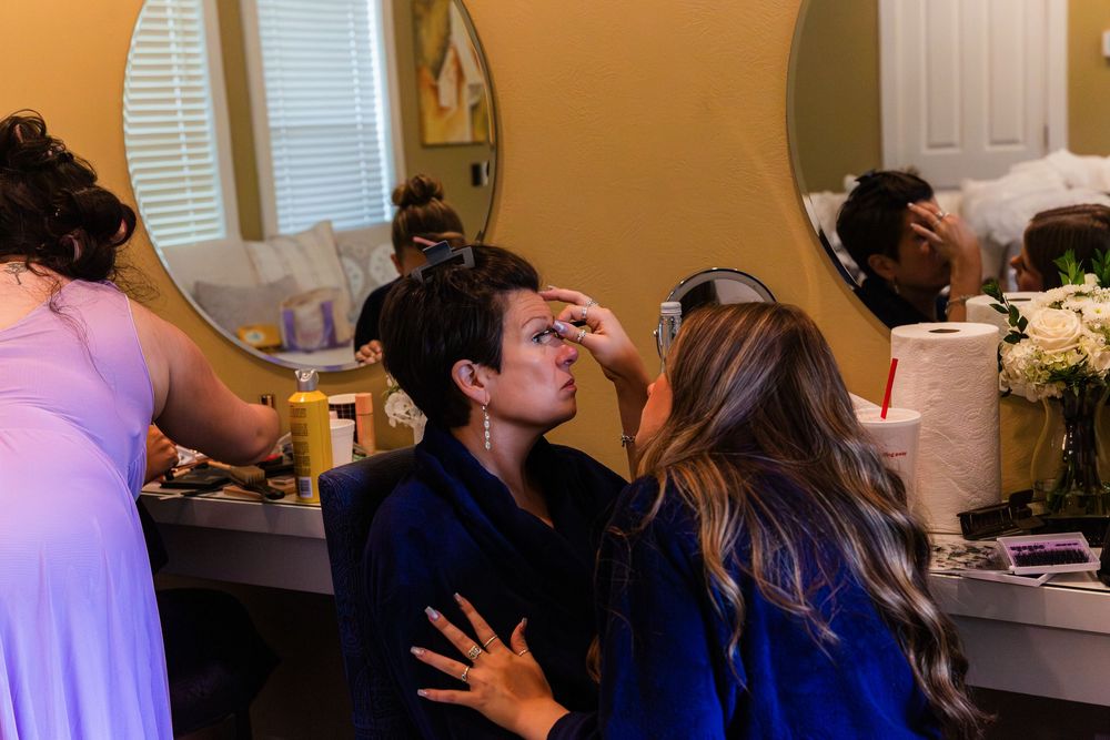 Bride getting makeup applied by artist while bridesmaid assists in elegant dressing room before wedding.