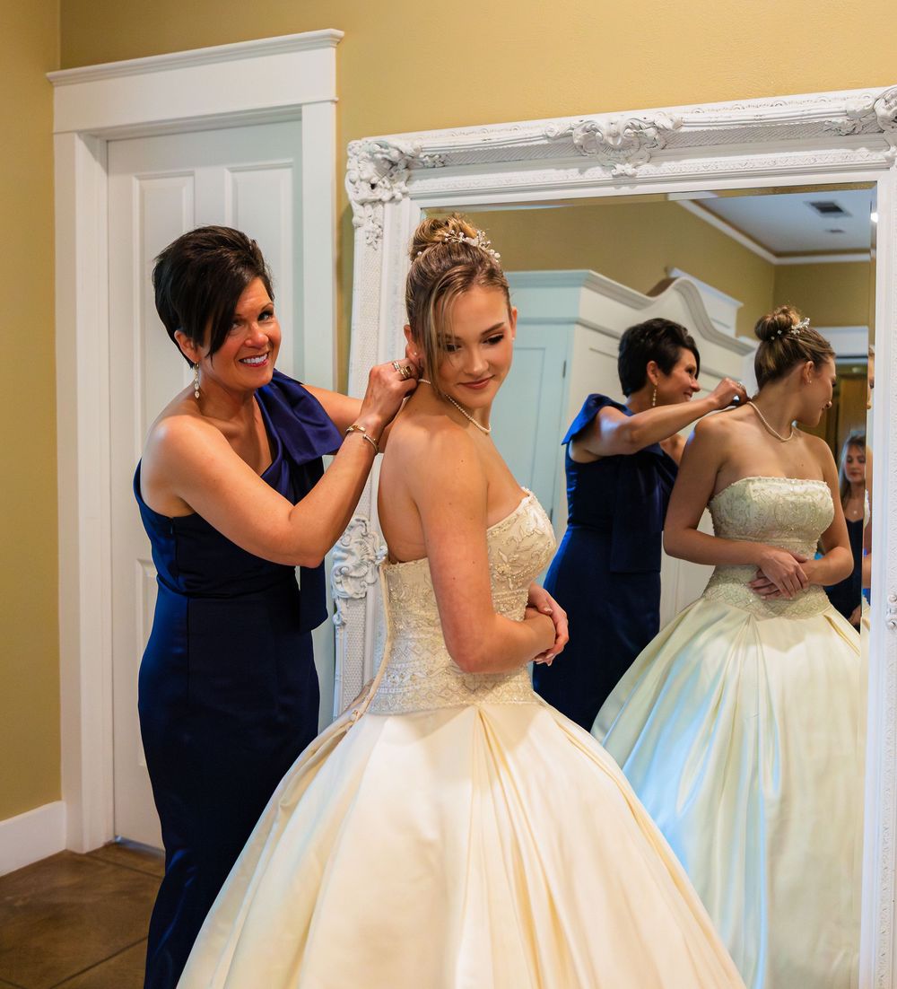 A woman in blue dress helps bride in white ball gown with necklace, reflected in ornate mirror.