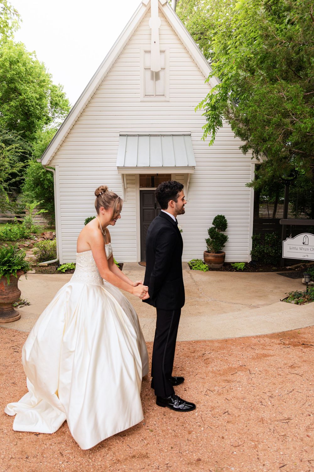 Bride and groom hold hands outside a white chapel surrounded by lush greenery on their wedding day.