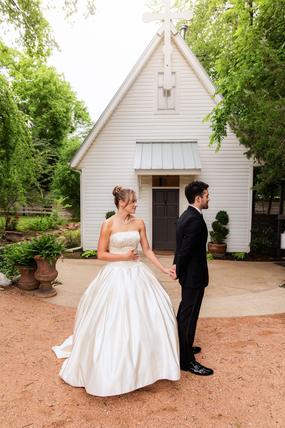 Bride and groom hold hands outside a white chapel with a cross, surrounded by lush green trees on their wedding day.