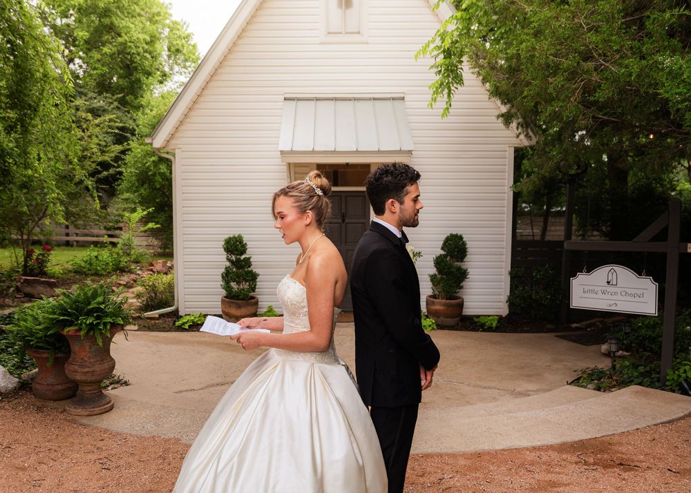 Bride in white ball gown and groom in black tuxedo reading letters back-to-back outside a white chapel.