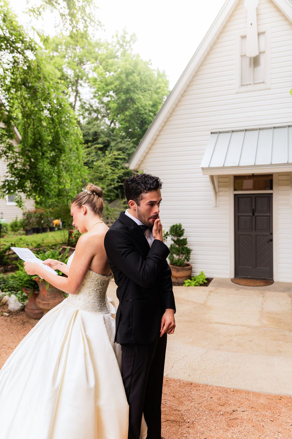 Bride and groom share emotional first look moment outside white chapel, groom covering mouth in tears.