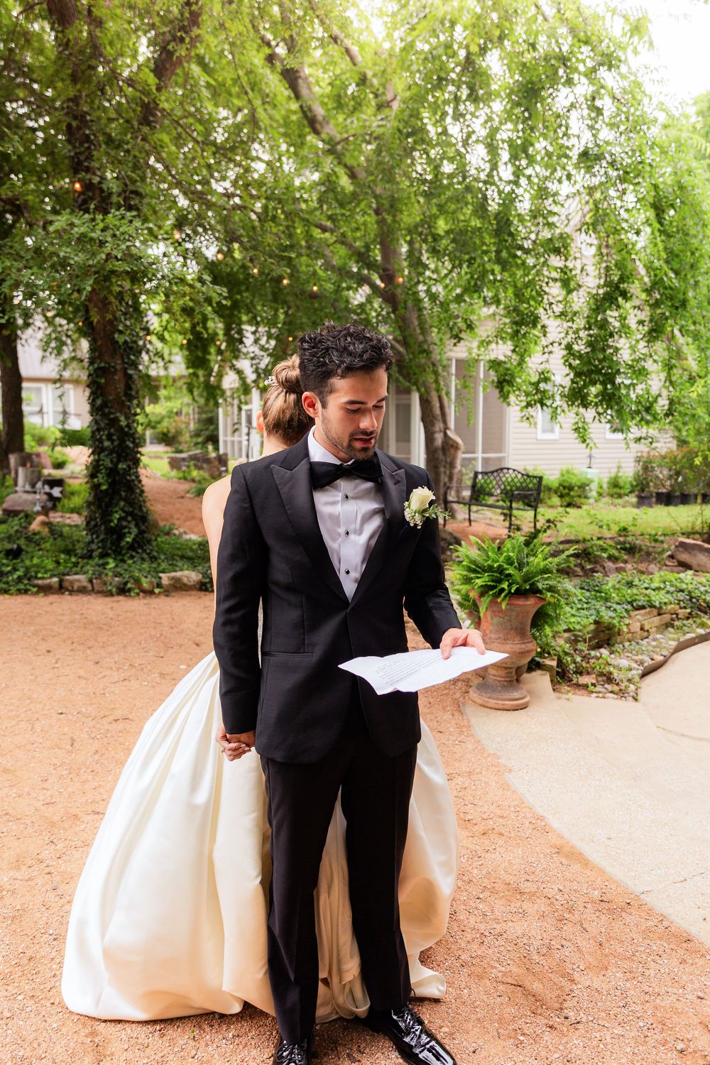 Groom in black tuxedo reads vows letter while bride in white gown stands behind him in a garden setting.