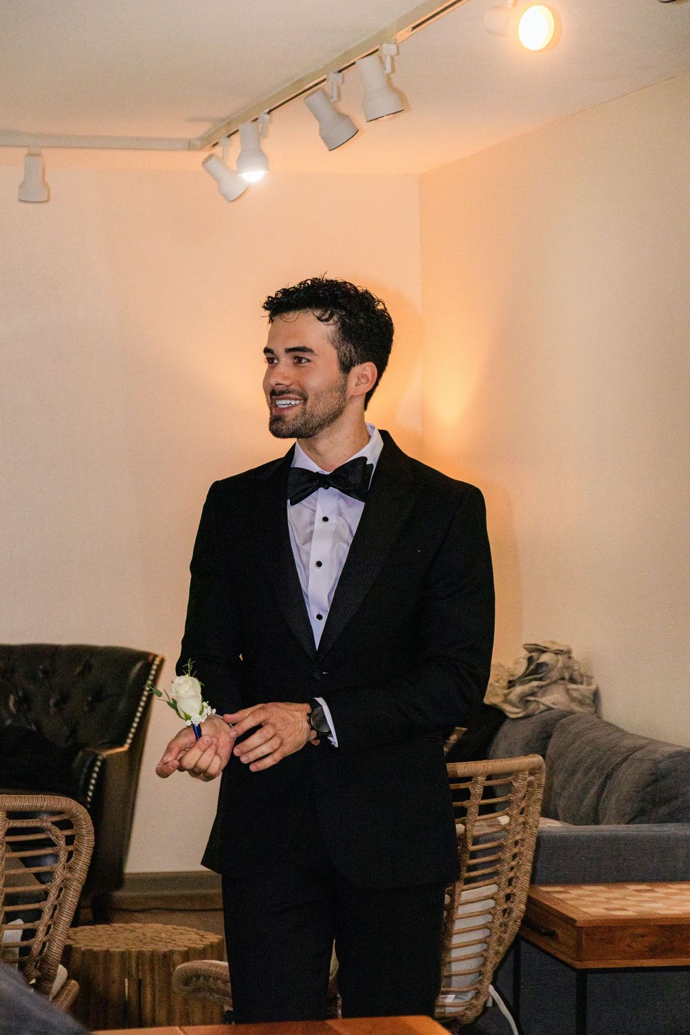 Handsome groom in black tuxedo with boutonniere smiles in warmly lit room before wedding.