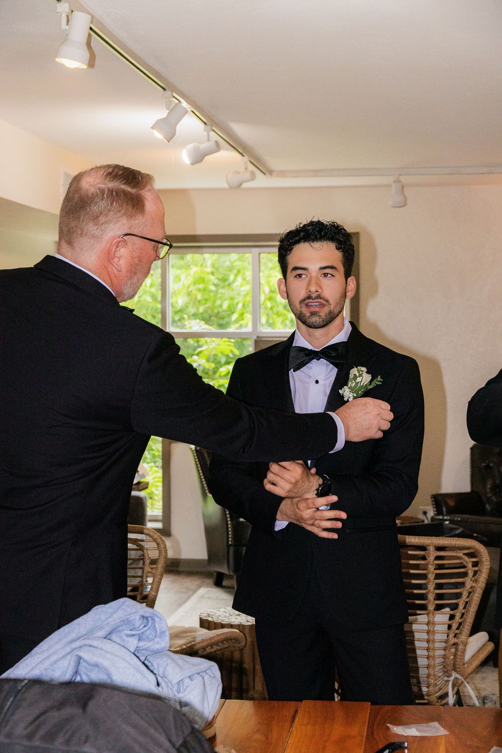 Older man pins boutonniere on groom's black tuxedo lapel before wedding ceremony.
