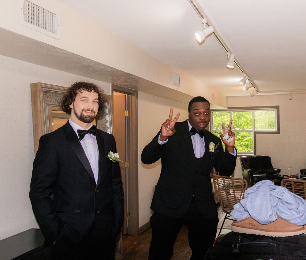 Two groomsmen in black tuxedos pose playfully in getting-ready room before wedding ceremony.