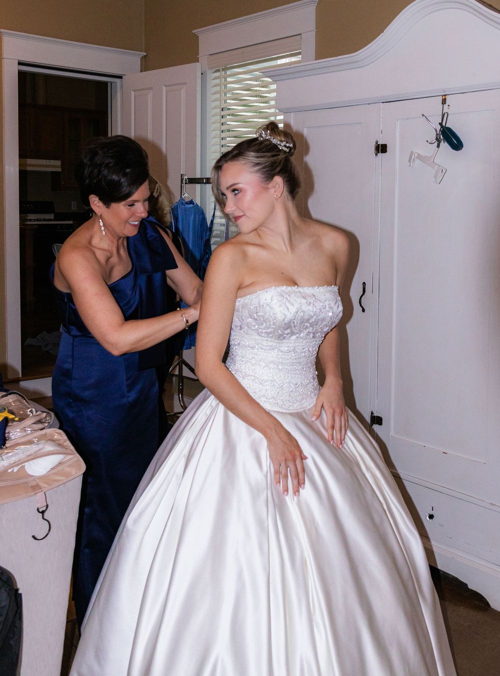 Mother helping bride into strapless white ballgown wedding dress in elegant dressing room.