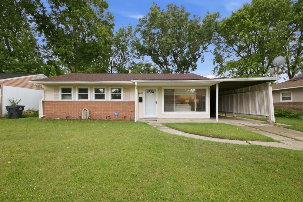 Brick ranch-style home with carport, white front door, manicured lawn, and mature trees in a suburban neighborhood.