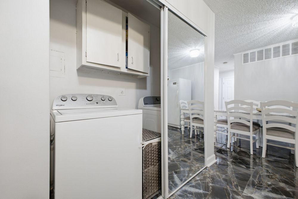 Laundry area with white washer, upper cabinet, and sliding mirror doors opening to a dining room with marble floors.