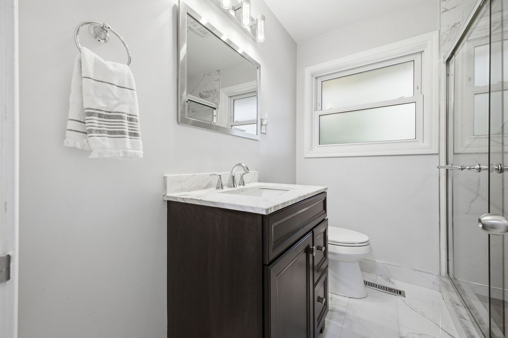 Modern bathroom with dark wood vanity, white marble floors, frameless glass shower, and bright natural light from frosted window.