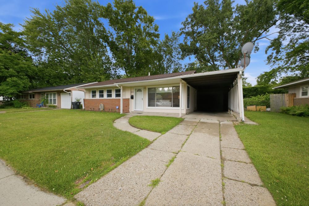 Ranch-style brick home with attached carport, large front lawn, and mature trees on a sunny day.