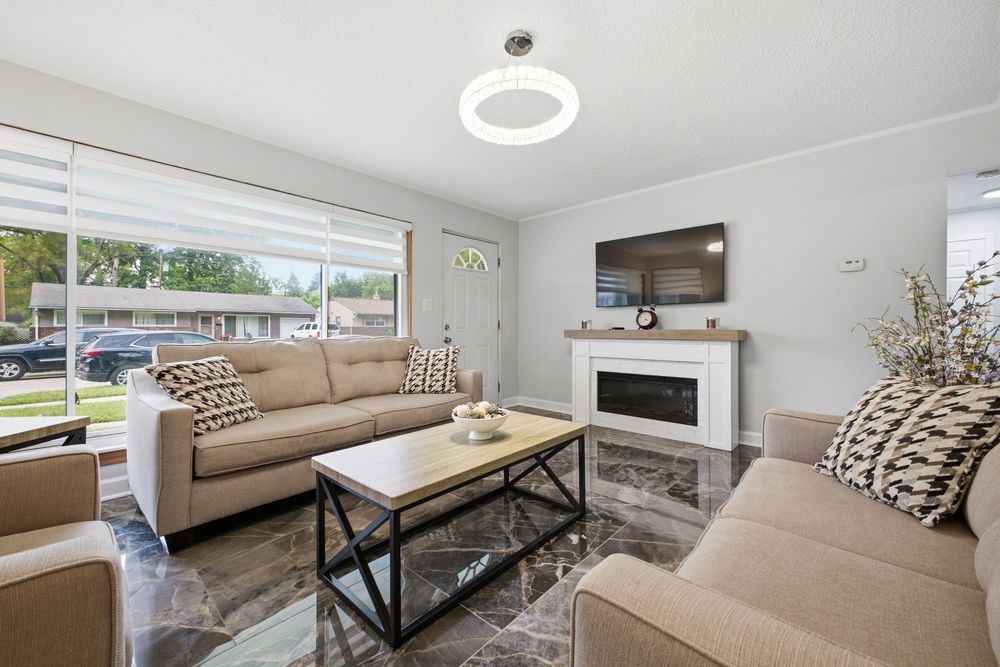 Modern living room with beige sofas, marble floors, electric fireplace, wall-mounted TV, and large windows with natural light.
