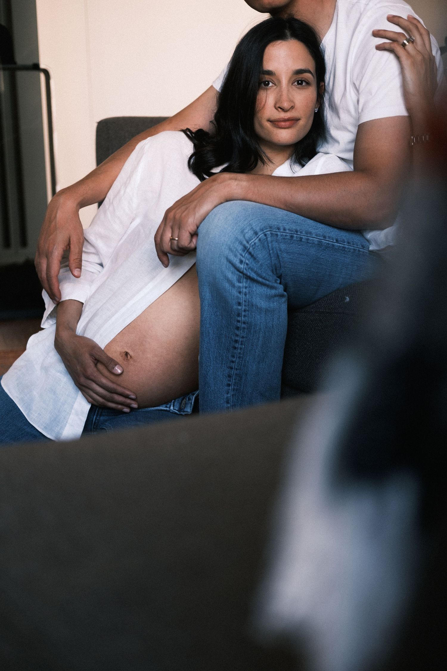 Pregnant woman in white shirt and jeans sitting between partner's legs on couch, cradling baby bump.
