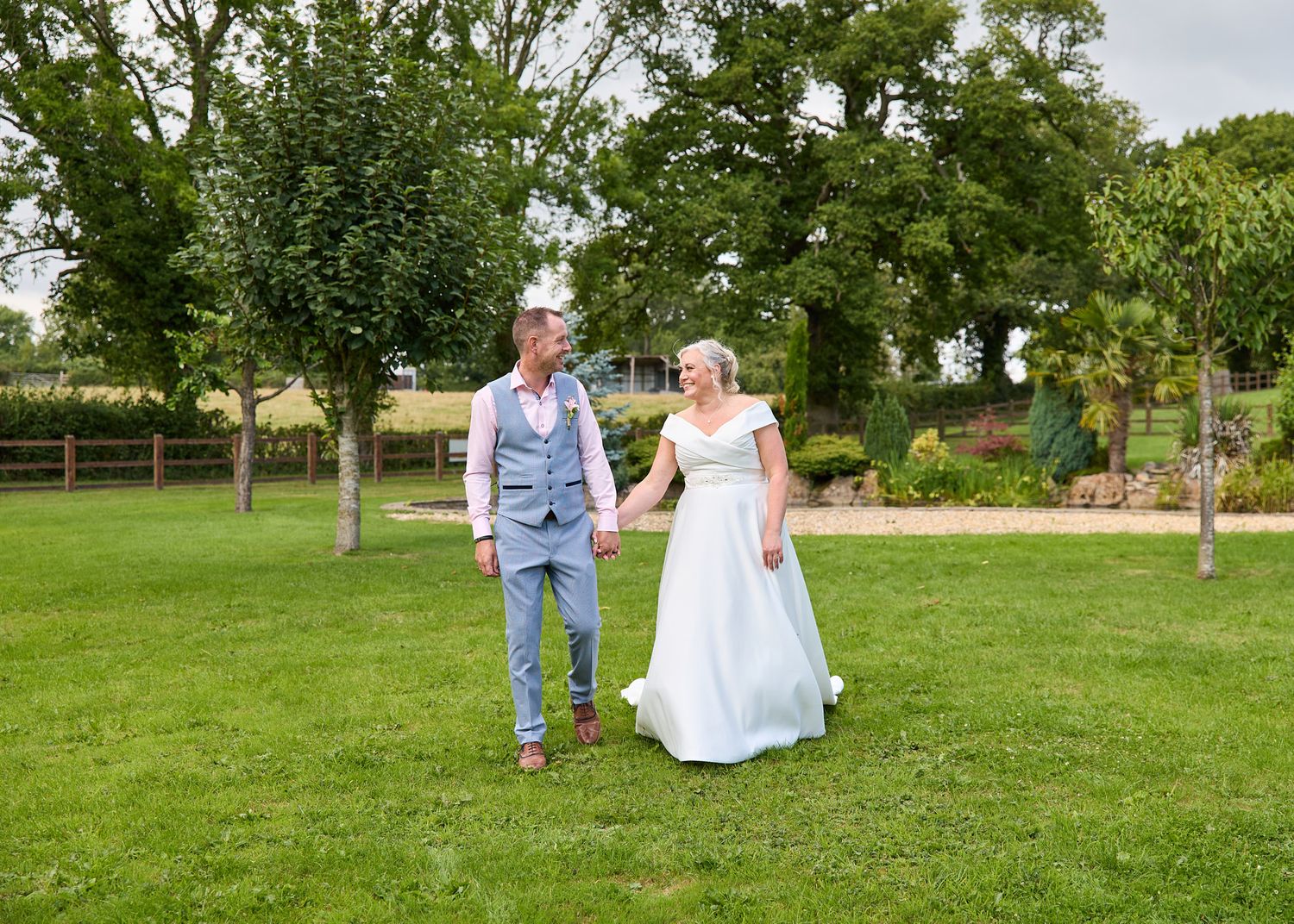 Bride and groom holding hands walking across green lawn at outdoor summer wedding venue.