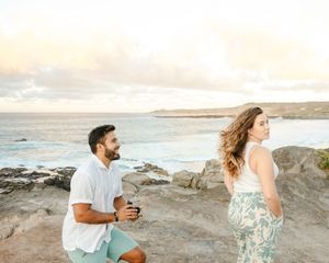 Man kneels on rocky coastal cliff preparing to propose to woman at sunset.