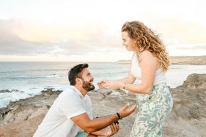 Couple shares romantic engagement moment on rocky Hawaii beach at golden sunset.