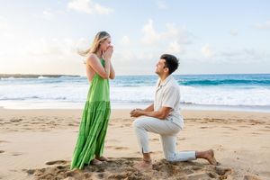 Man proposing on one knee to woman in green dress on sandy beach at sunrise.